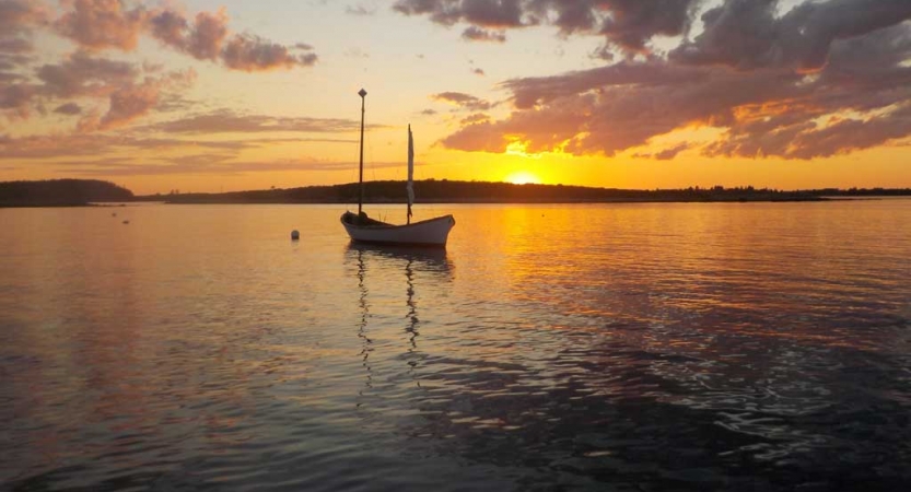 A sailboat floats on calm water, reflecting the orange and yellow sunset. 